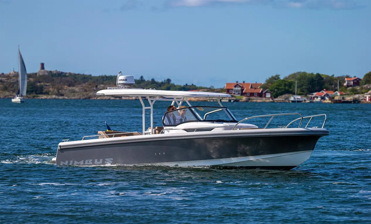 A Nimbus boat navigates blue waters, with passengers aboard and coastal houses visible in the background under a clear sky.